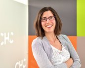 Eine Frau mit schulterlangen Haaren und Brille lächelt, während sie mit verschränkten Armen vor einer farbigen Wand steht. Die Wand hat orange, grüne und graue Elemente.